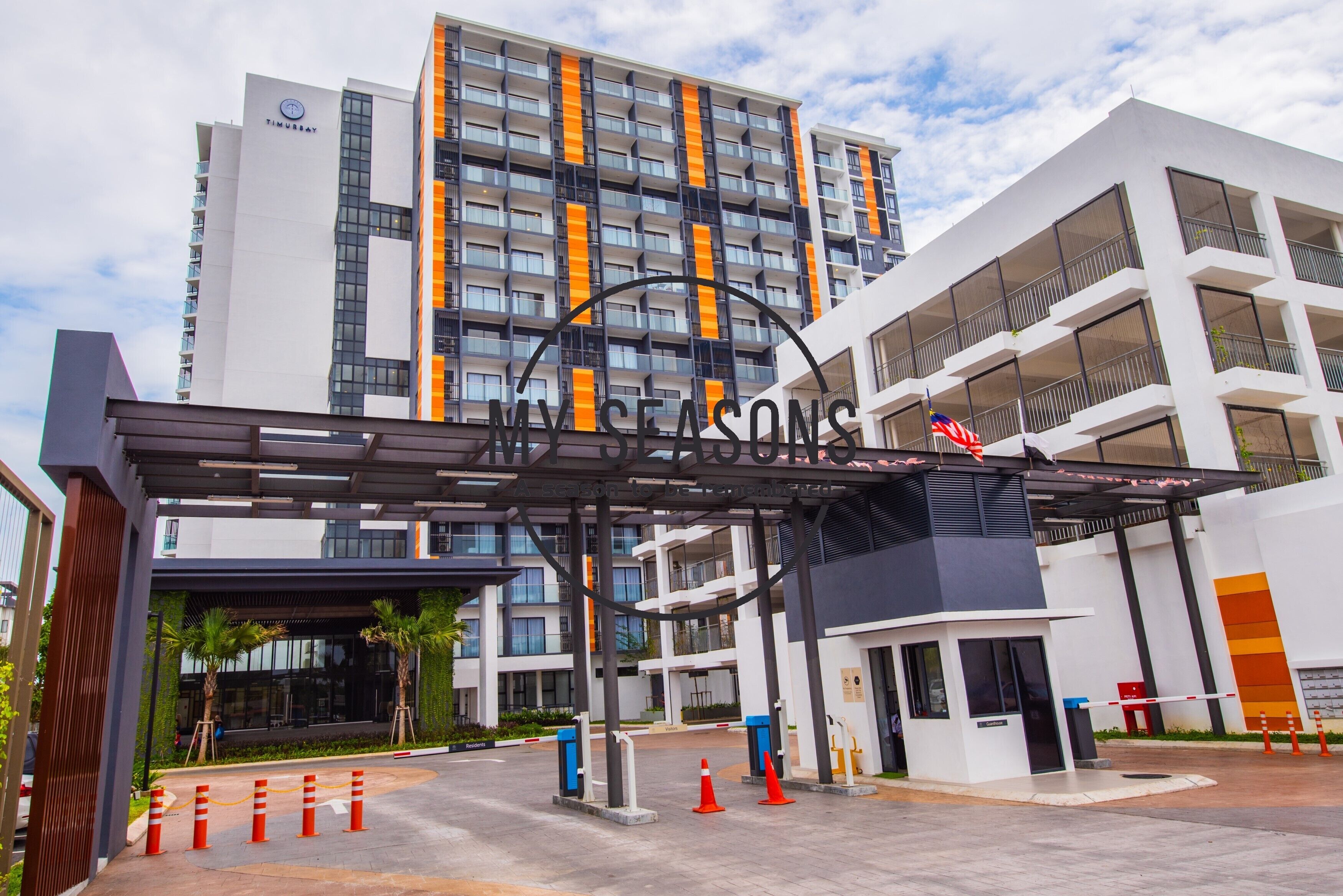 A modern hotel entrance with a prominent sign, a gated driveway, and a multi-story building with balconies.