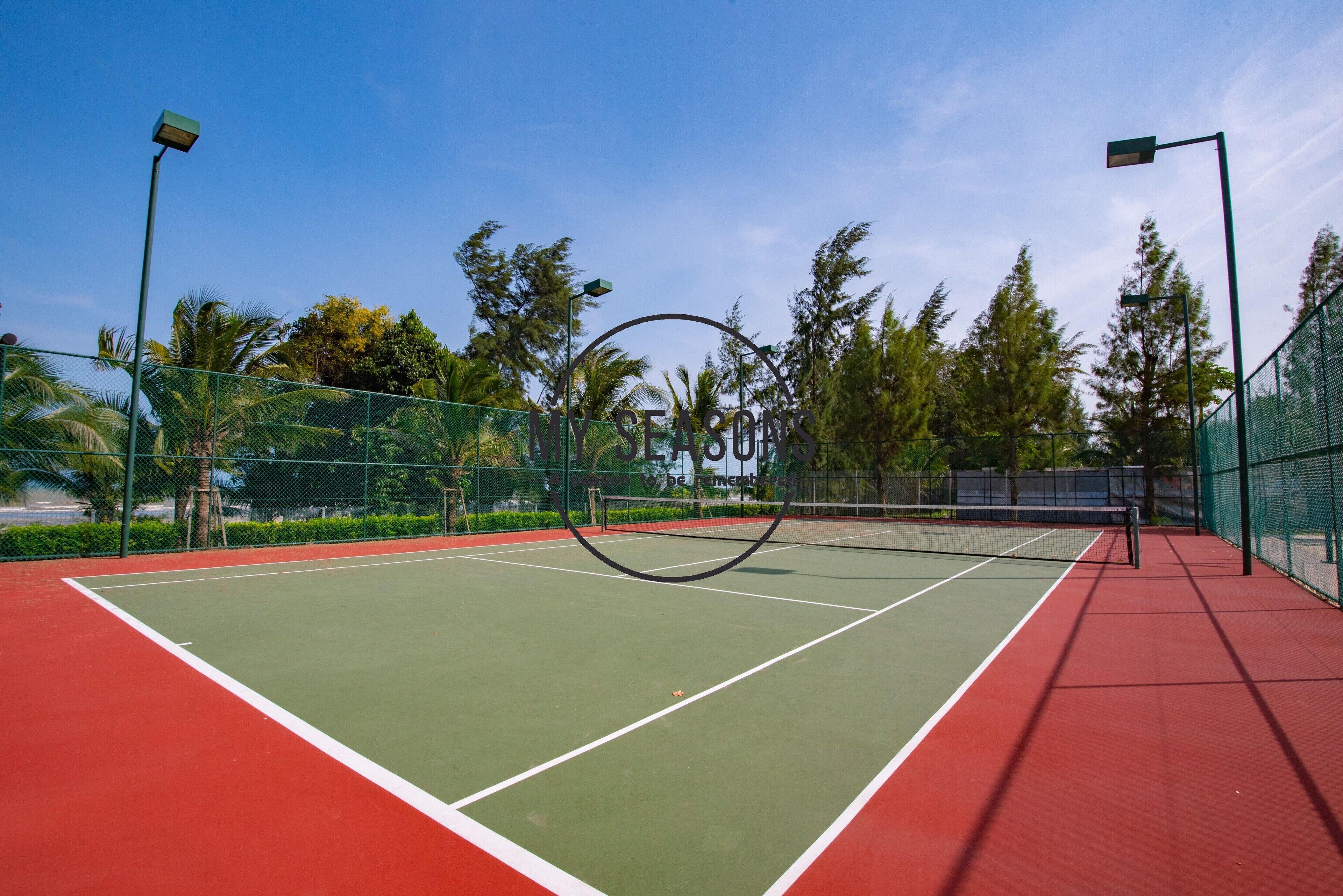 A tennis court with a net, surrounded by a green fence and palm trees.