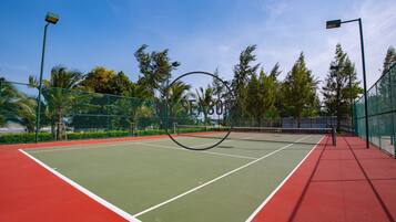 A tennis court with a net, surrounded by a green fence and palm trees.