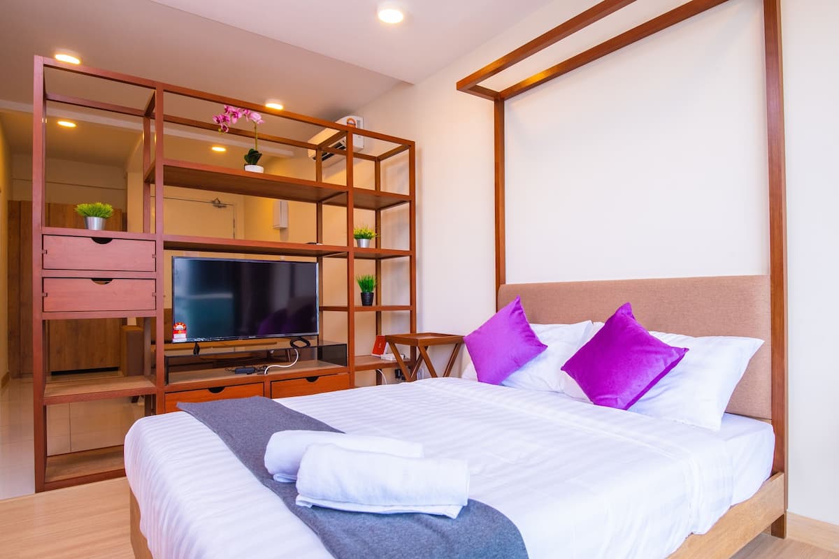 A hotel room with a bed, a TV, a wooden desk, and a shelving unit with plants.