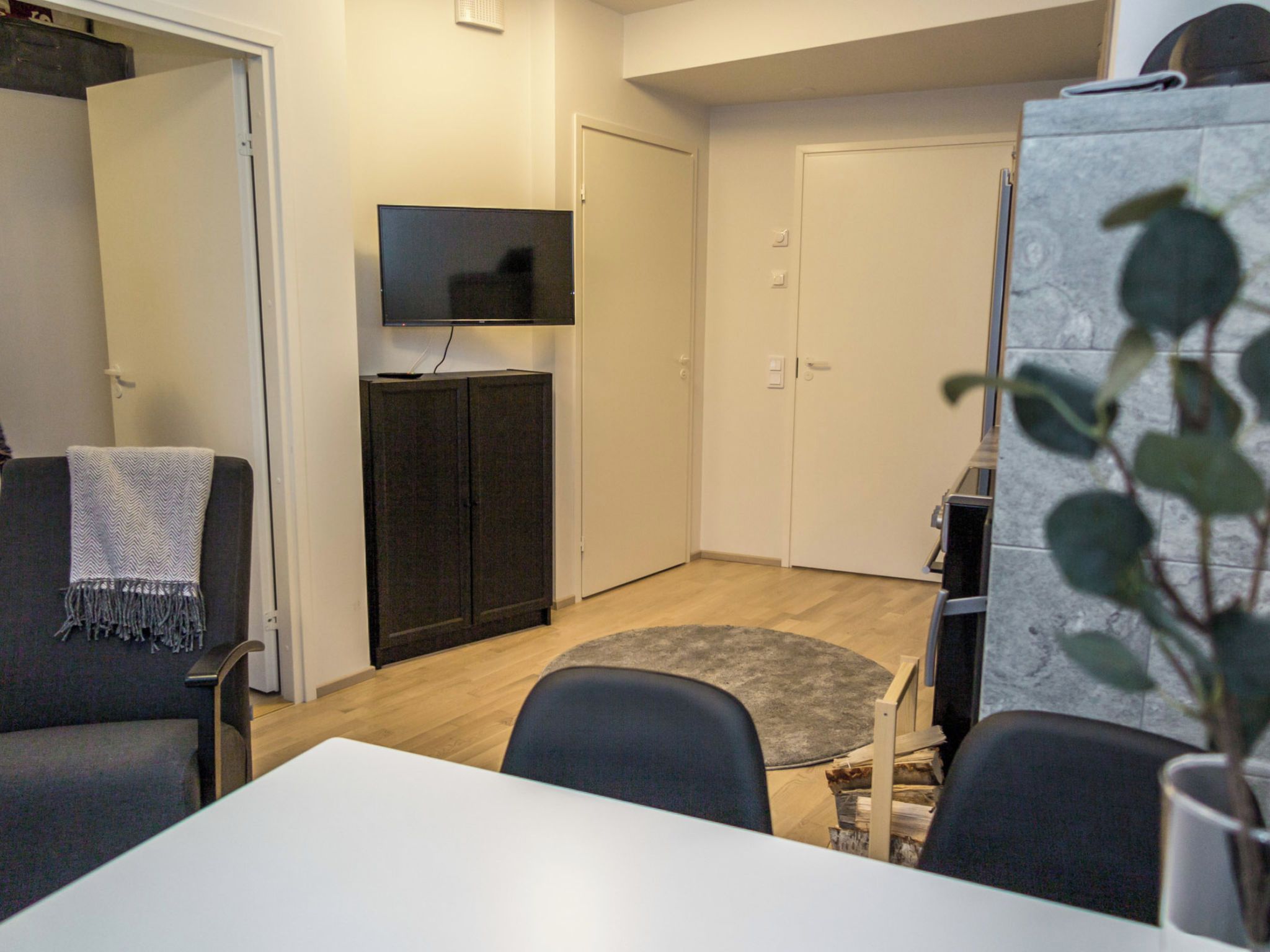 Un espacio habitable pequeño con un televisor montado en la pared, un mueble auxiliar, una mesa de comedor con sillas y una planta en primer plano.