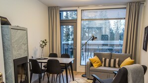 A modern living room with a fireplace, a dining table with chairs, a sofa with cushions, and large windows with a view of trees.
