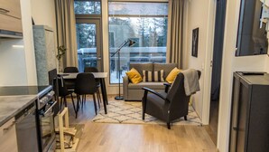 A modern kitchen with a built-in oven, dining area with a table and chairs, a sofa with cushions, and a large window with a view of trees.