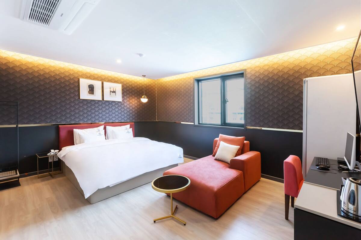 Ein modernes Hotelzimmer mit einem großen Bett, einem roten Sofa, einem kleinen runden Tisch und einem Schreibtisch mit Computer und Wasserkocher.