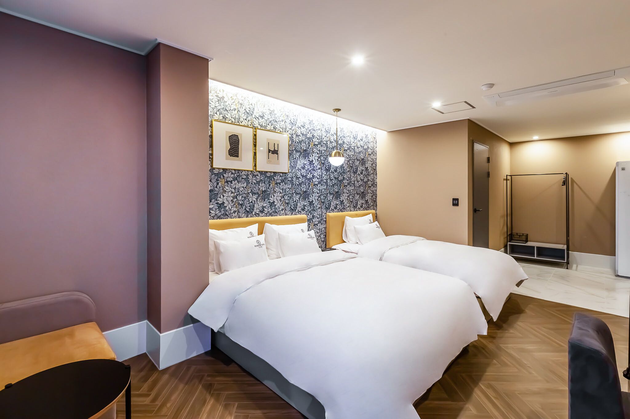 A hotel room with two beds, a wooden floor, a bench, a wall with framed pictures, and a shower area.
