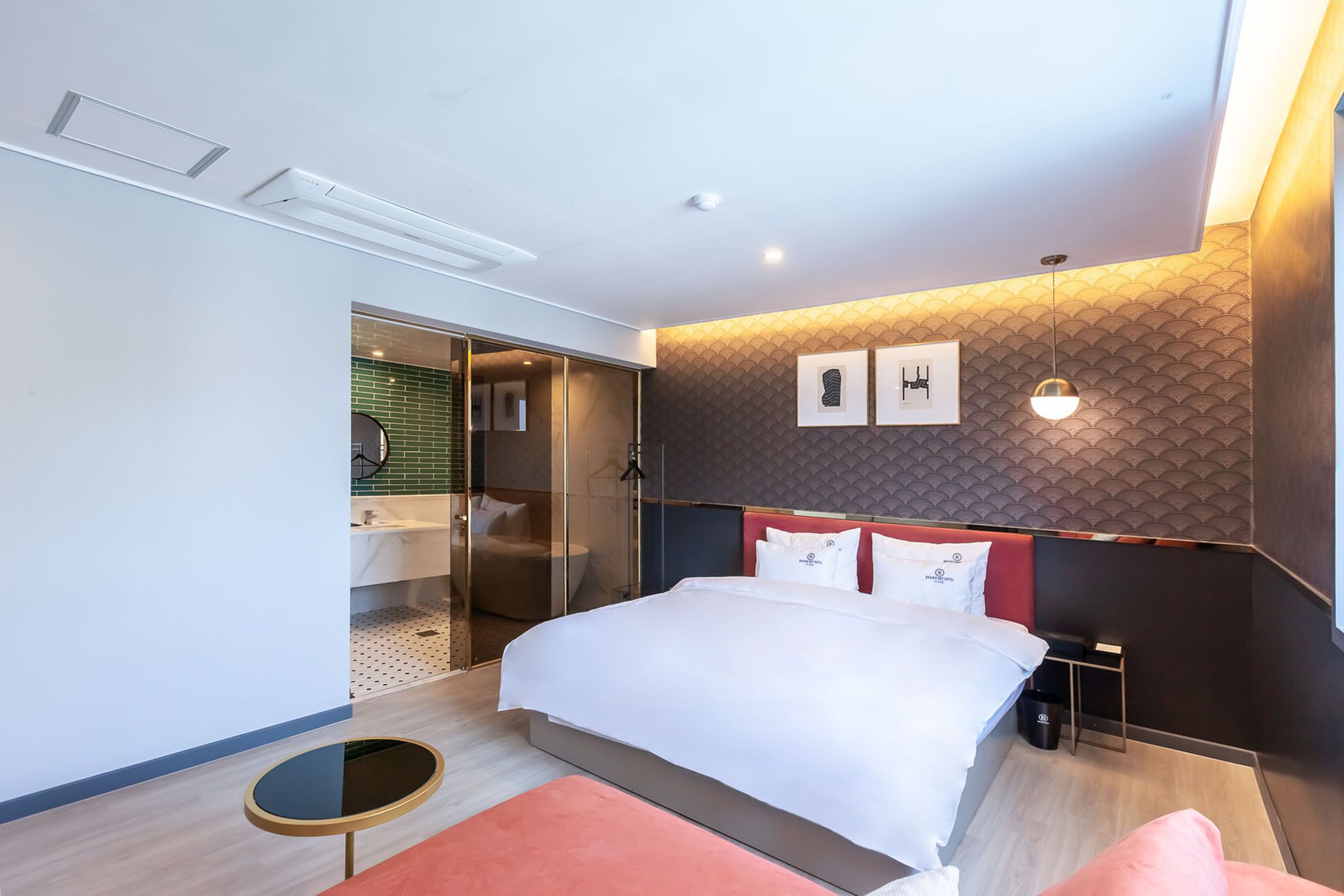 Una habitación de hotel moderna con una cama grande, una pequeña mesa redonda, una zona de ducha con cerramiento de vidrio y una pared de acento roja.