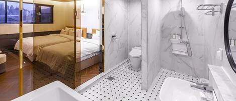 A modern hotel room with a glass partition separating the bedroom and bathroom. The bathroom features a shower, toilet, and a sink with a mirror.