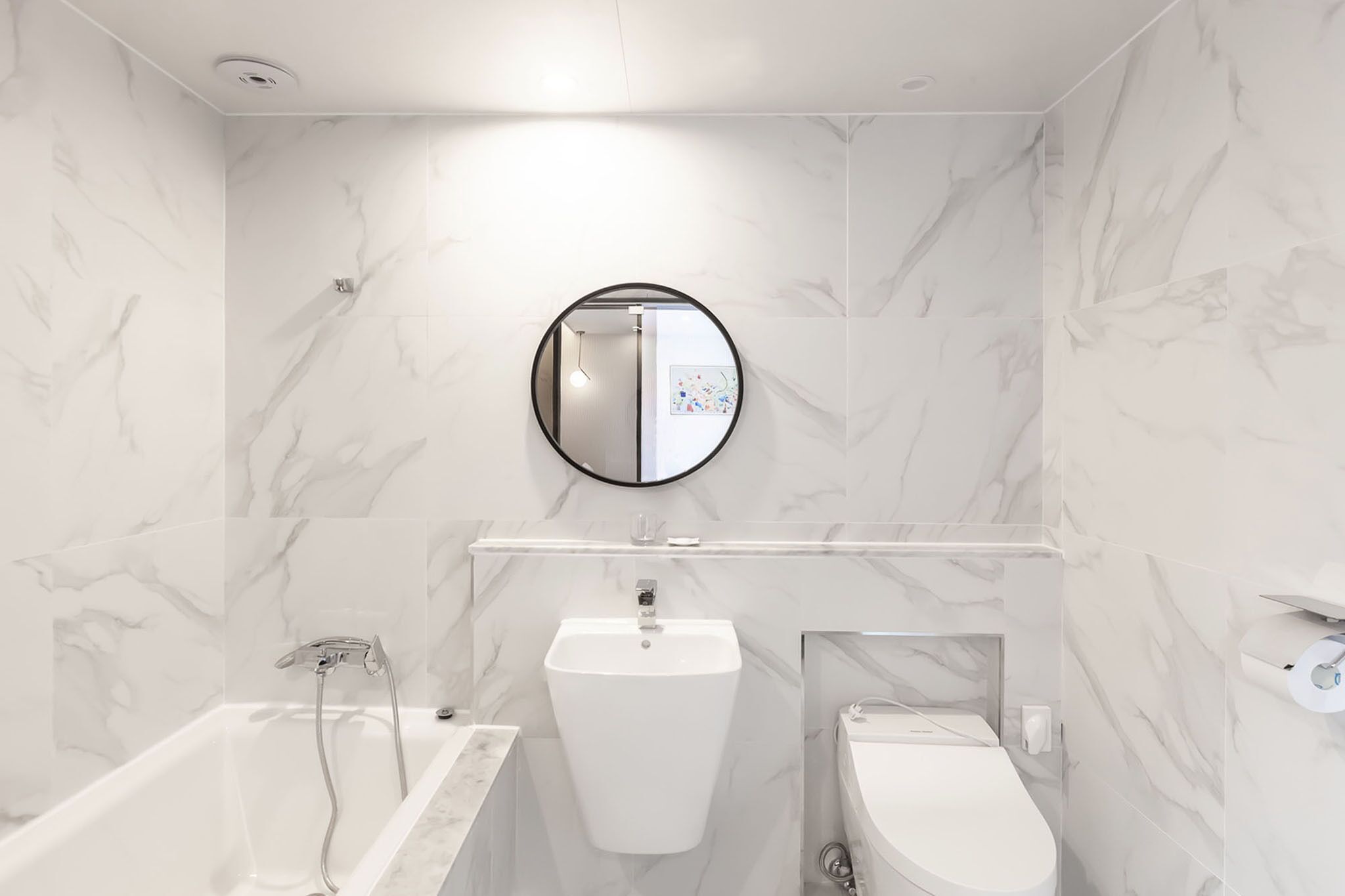 A modern bathroom with a marble wall, a round mirror, a pedestal sink, a bathtub, and a toilet.