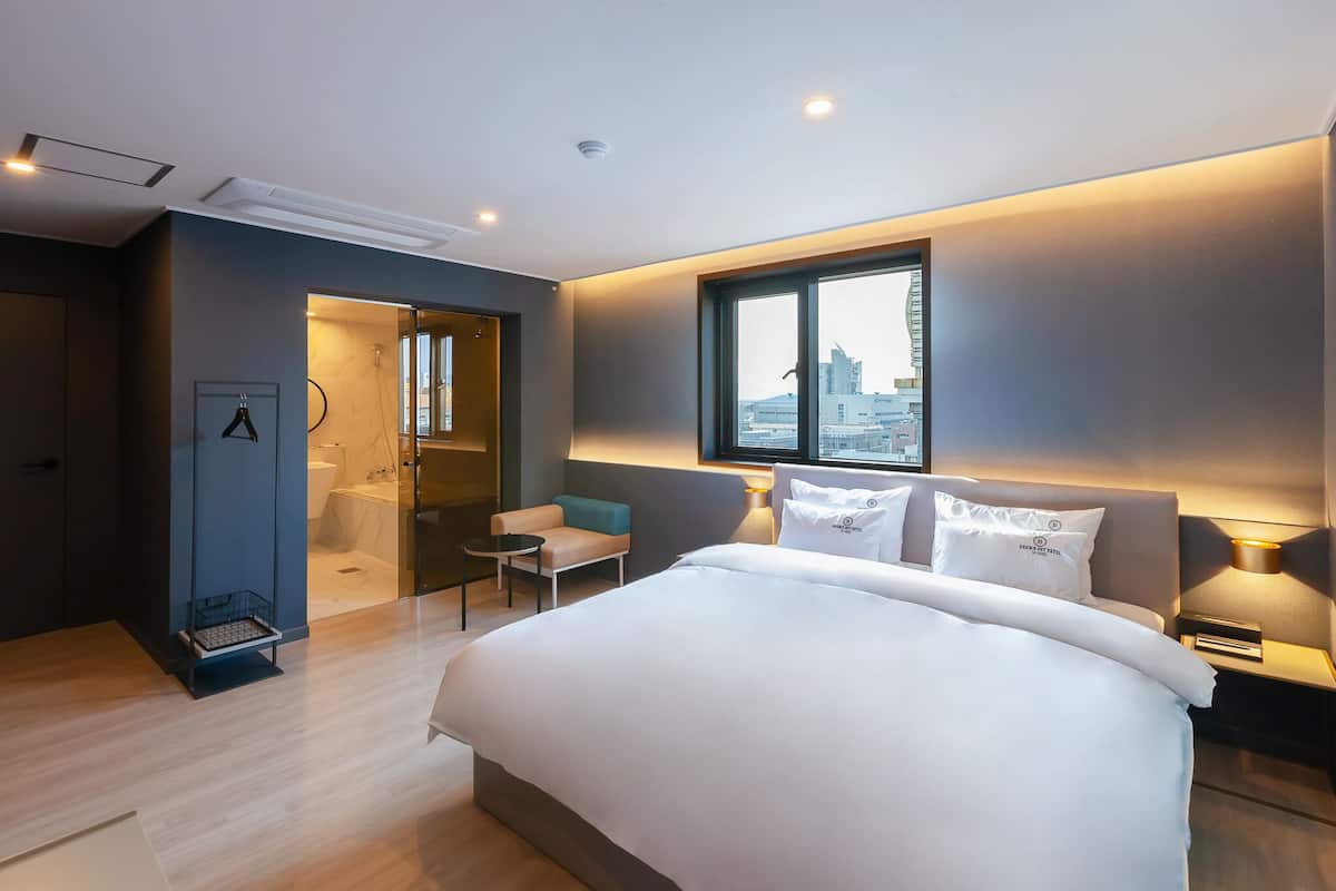 Ein modernes Hotelzimmer mit einem großen Bett, einer Sitzecke, einem Fenster mit Blick auf die Stadt und einem separaten Badezimmer.