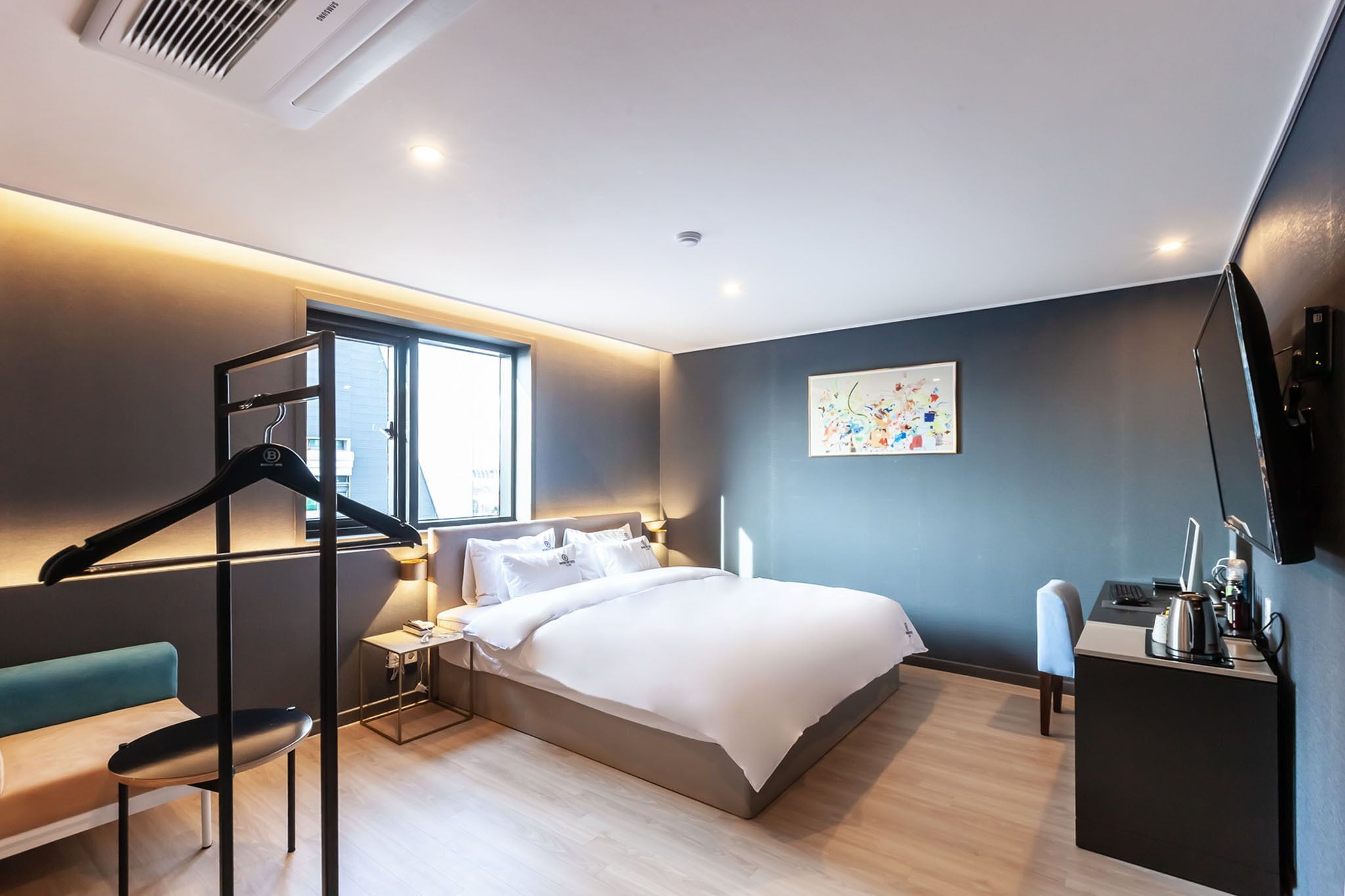 A modern hotel room with a large bed, a desk, and a chair. There is a window providing natural light and a wall-mounted TV.