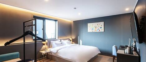 A modern hotel room with a large bed, a desk, and a chair. There is a window providing natural light and a wall-mounted TV.