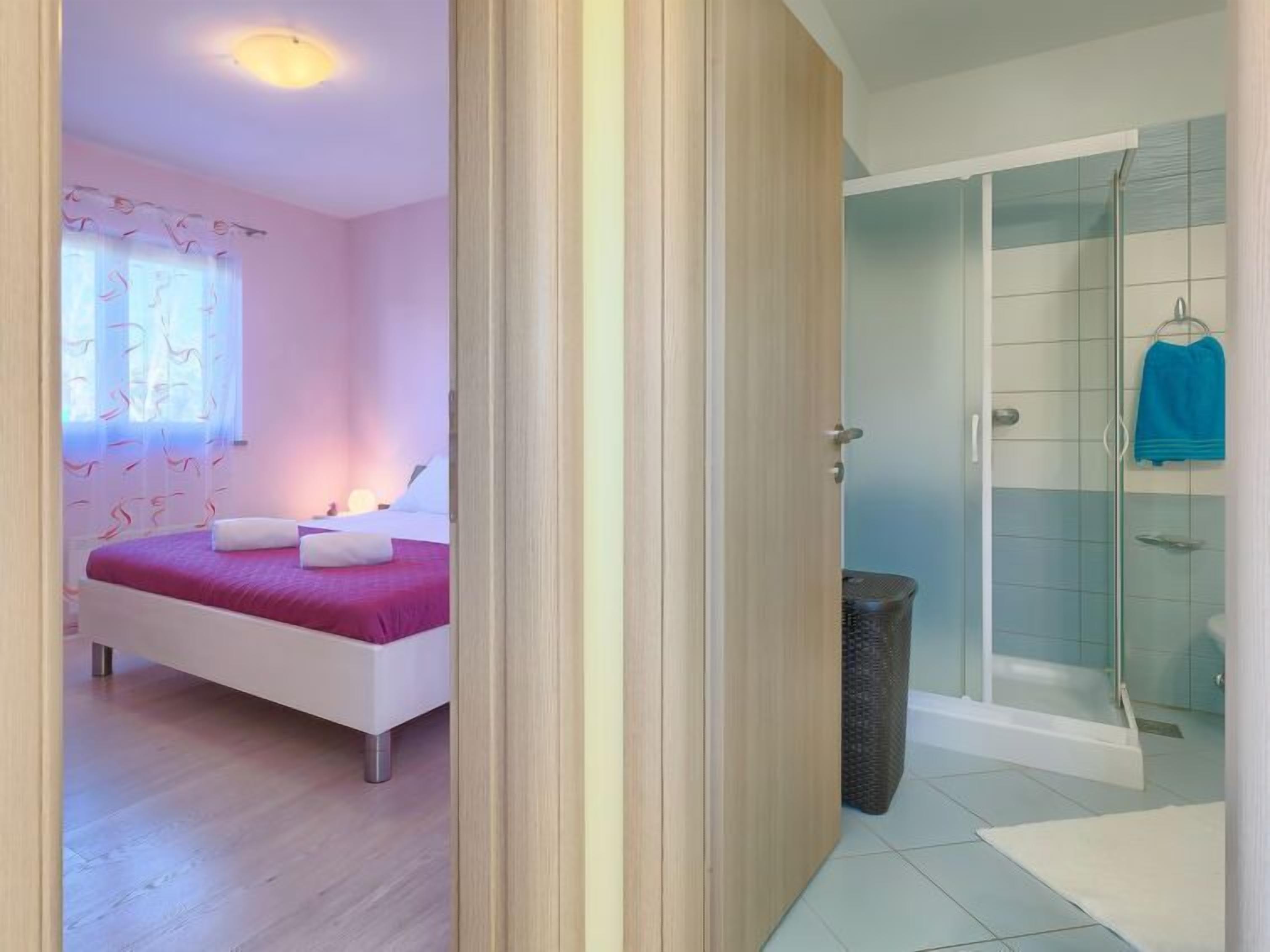 A bedroom with a bed, a bathroom with a shower, and a visible trash bin.