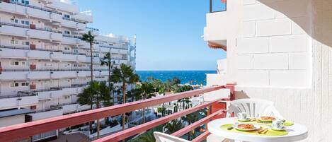 Un balcón con mesa lista para comer, con vista a un paisaje urbano costero con edificios y palmeras.