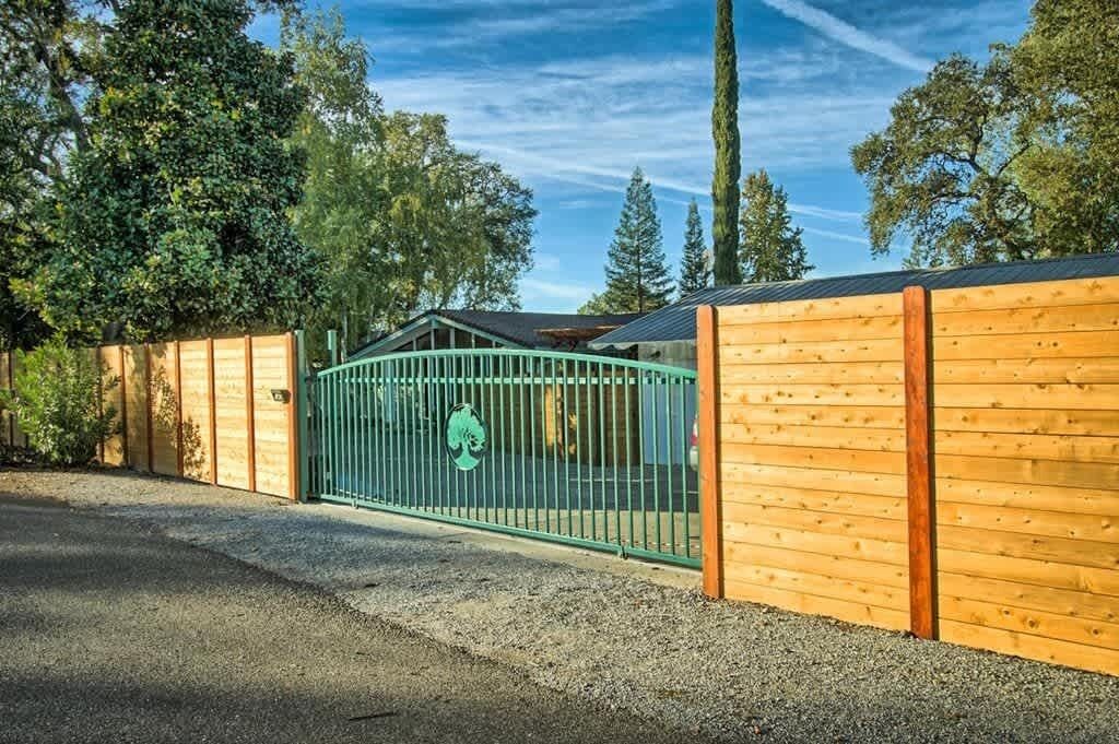 A gated wooden fence with a metal gate, a gravel driveway, and trees in the background.