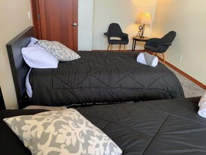 Una cama doble con cabecera oscura, dos mesitas de noche, una lámpara y dos sillas en una habitación de hotel.