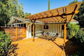 Una terraza de madera con una pérgola, un área cercada y un edificio con techo inclinado.