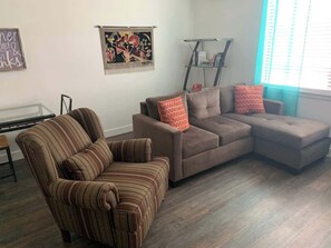Uma sala de estar com uma poltrona listrada, um sofá marrom e um sofá seccional com almofadas vermelhas. Há uma mesa de centro de vidro, uma estante e uma pintura na parede.