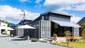 A modern building with a large shipping container structure, a covered outdoor seating area, and a visible power line tower in the background. - Cottage:Di Musica (Fujikawaguchiko)