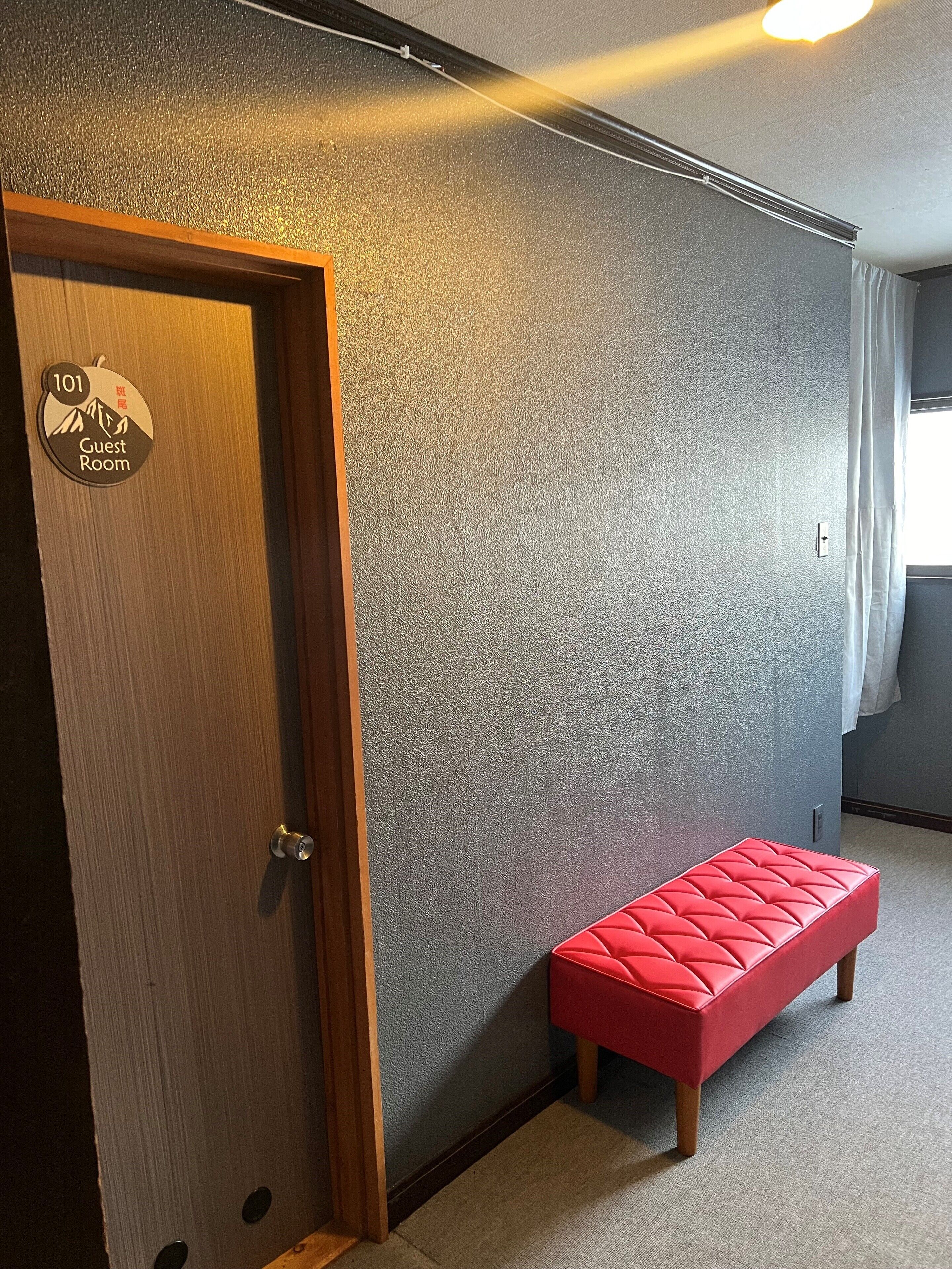 Quarto de hotel com parede cinza texturizada, porta de madeira com a placa '101 Guest Room' e um banco vermelho.