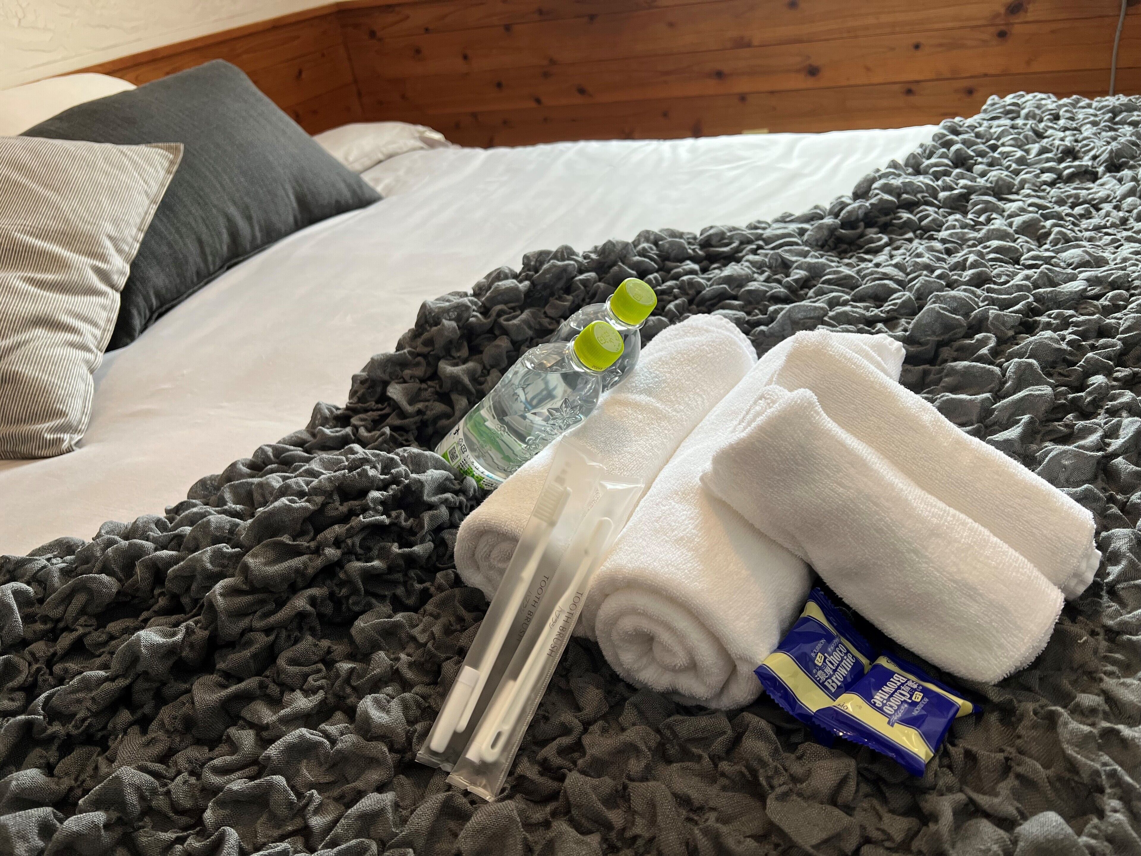 A neatly made bed with a grey textured blanket, two rolled white towels, a clear bottle with a green cap, and a pair of white slippers.