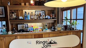 A wooden interior with a counter displaying 'Ringo Jam MADARAO' signage, shelves with bottles and snacks, and a large window with a view. - Ringo Jam Madarao (Myoko)