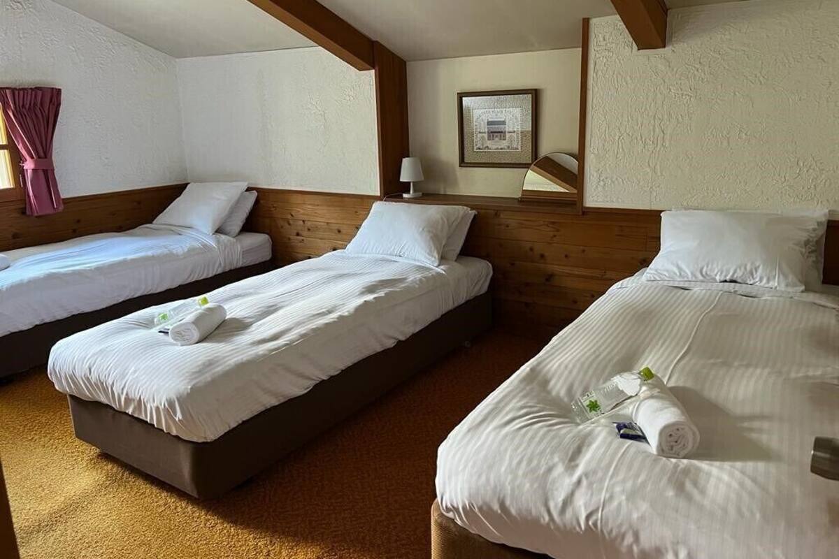 A hotel room with three single beds, wooden beams on the ceiling, a framed picture on the wall, and a small table with a lamp.