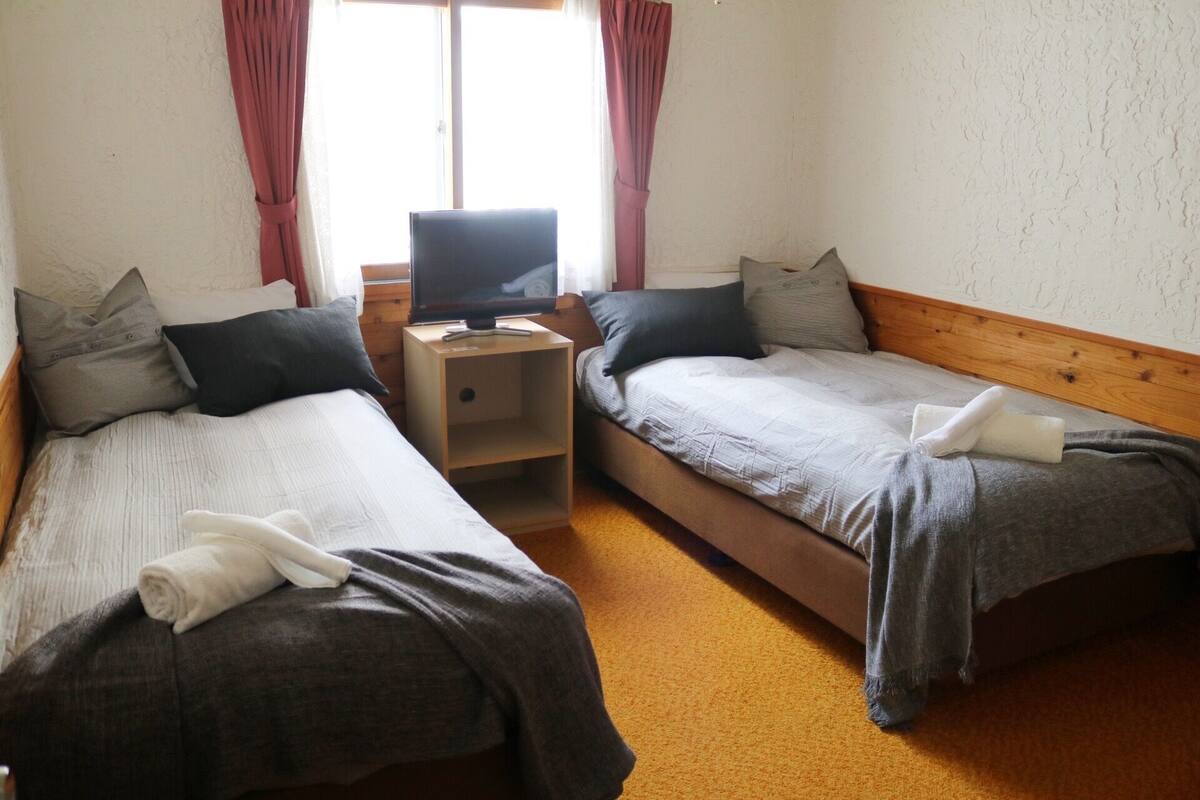 A room with two single beds, a TV on a wooden nightstand, and a window with curtains.