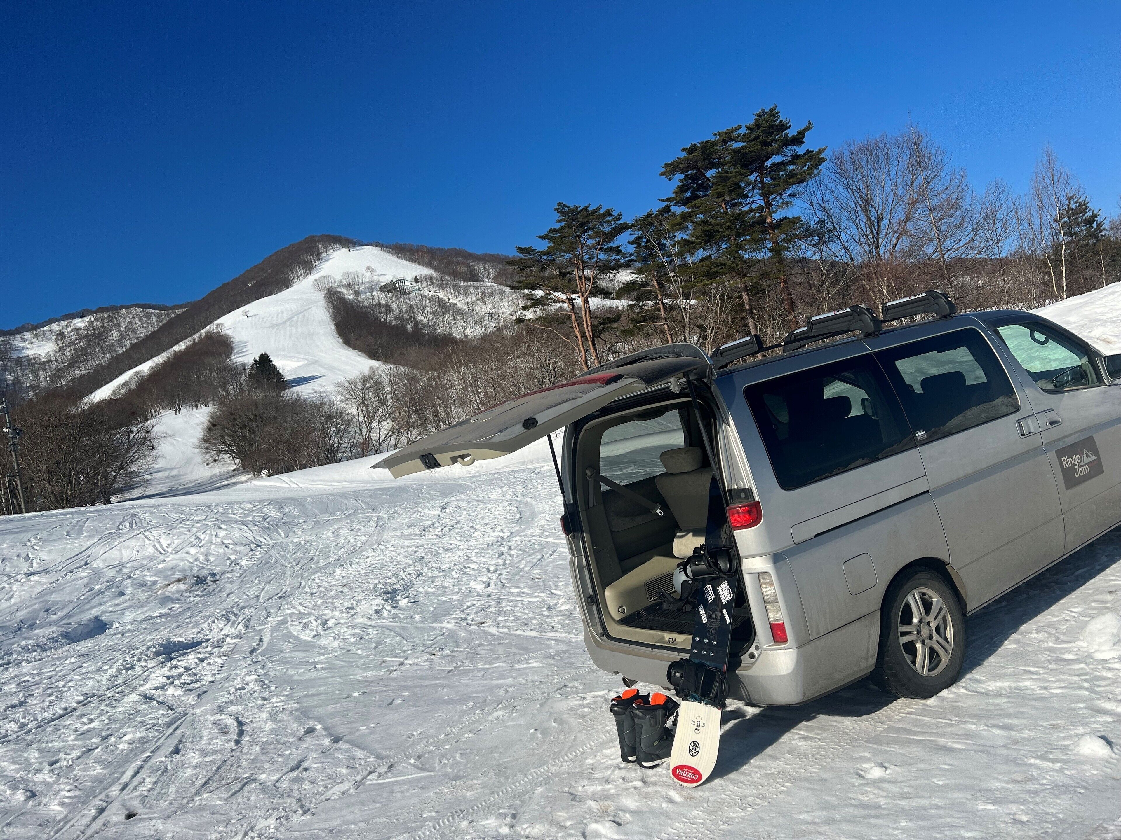 A silver van parked on a snowy slope with a snowboard attached to the back.