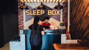 A reception desk with a 'SLEEP BOX' sign, a person standing behind it, and a wooden wall in the background. - Sleep Box Hostel Suratthani (Surat Thani)