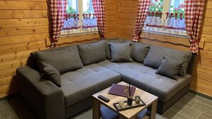 Un coin douillet avec un canapé d’angle gris, une petite table en bois sur laquelle repose une télécommande et des rideaux à carreaux.