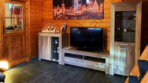 Un intérieur en bois avec un téléviseur posé sur un meuble en bois, une vitrine contenant des objets décoratifs et un tableau représentant un bâtiment illuminé par des feux d’artifice.