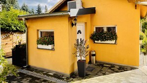 Un petit bâtiment jaune avec deux fenêtres, dont une ornée d’une jardinière et l’autre d’une plante suspendue.