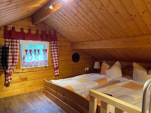 A wooden room with a sloped ceiling, a bed with a quilt, a window with curtains, and a lamp.