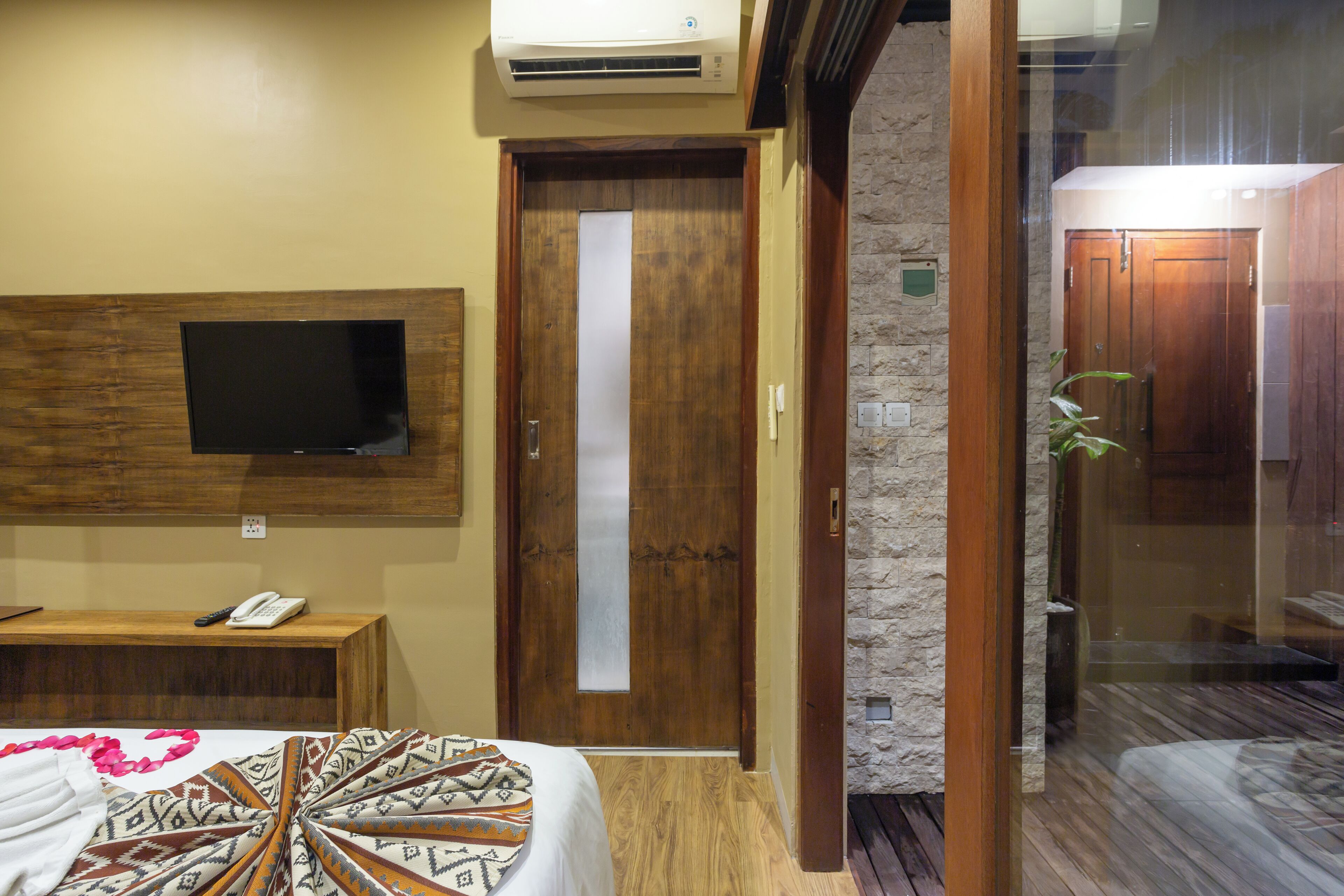 A hotel room with a bed, a TV mounted on the wall, a wooden desk, a telephone, and a door with a frosted glass panel.