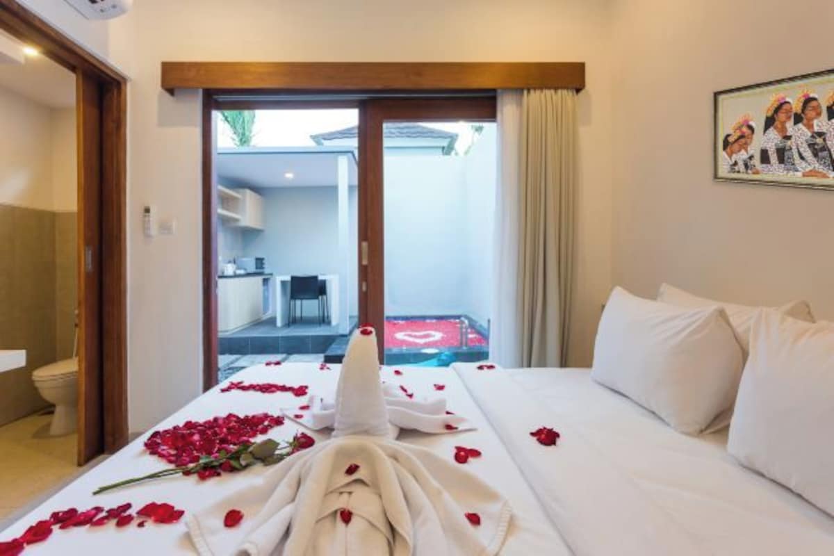 A hotel room with a bed covered in rose petals and a bouquet, a sliding glass door leading to a balcony, and a painting on the wall.