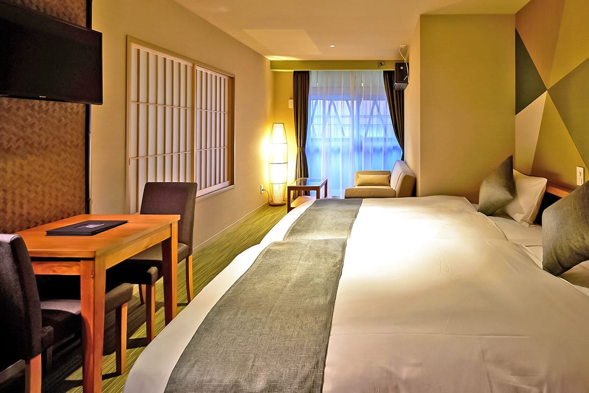 A hotel room with a large bed, a desk, a chair, a TV, and a view of the outside through a window.