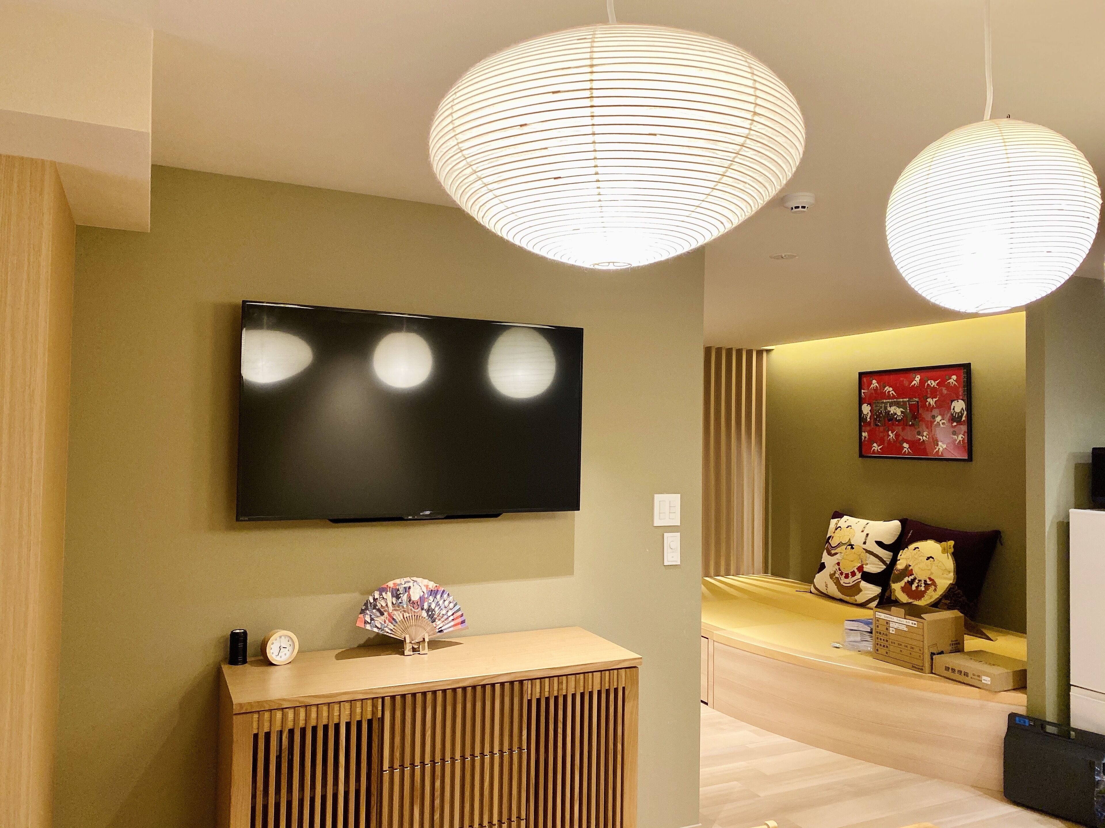 A modern living room with a flat-screen TV mounted on the wall, a wooden console table with a fan, and a sofa with decorative pillows.