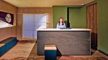 Un mostrador de recepción con una persona sentada detrás, un panel divisor de listones de madera, una pared verde y una ventana con persianas.