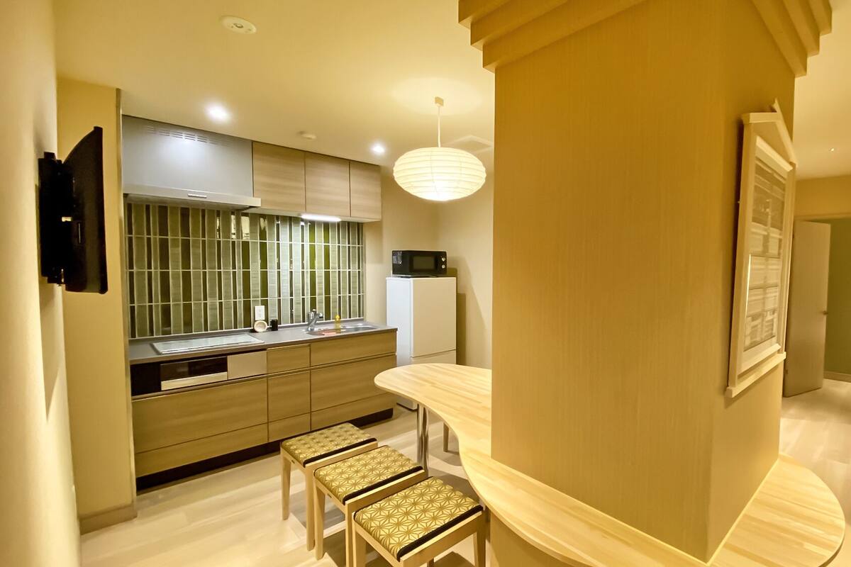 A modern kitchen with a built-in microwave, a sink, and a window with blinds. There are two stools and a pendant light.