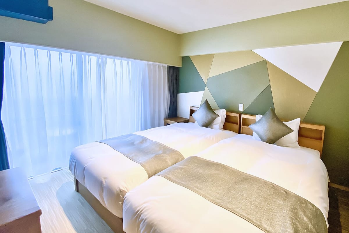 A hotel room with two beds, a large window with curtains, and a geometric wall design.