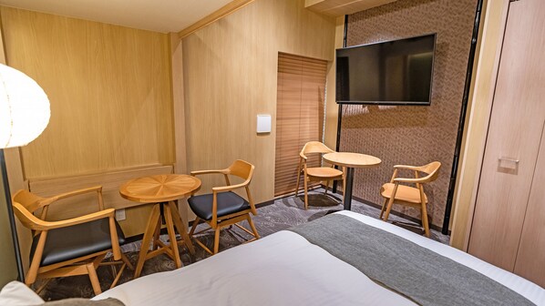 Habitación de hotel con cama, dos sillas de madera, una mesita redonda, televisor montado en la pared y un armario de madera.
