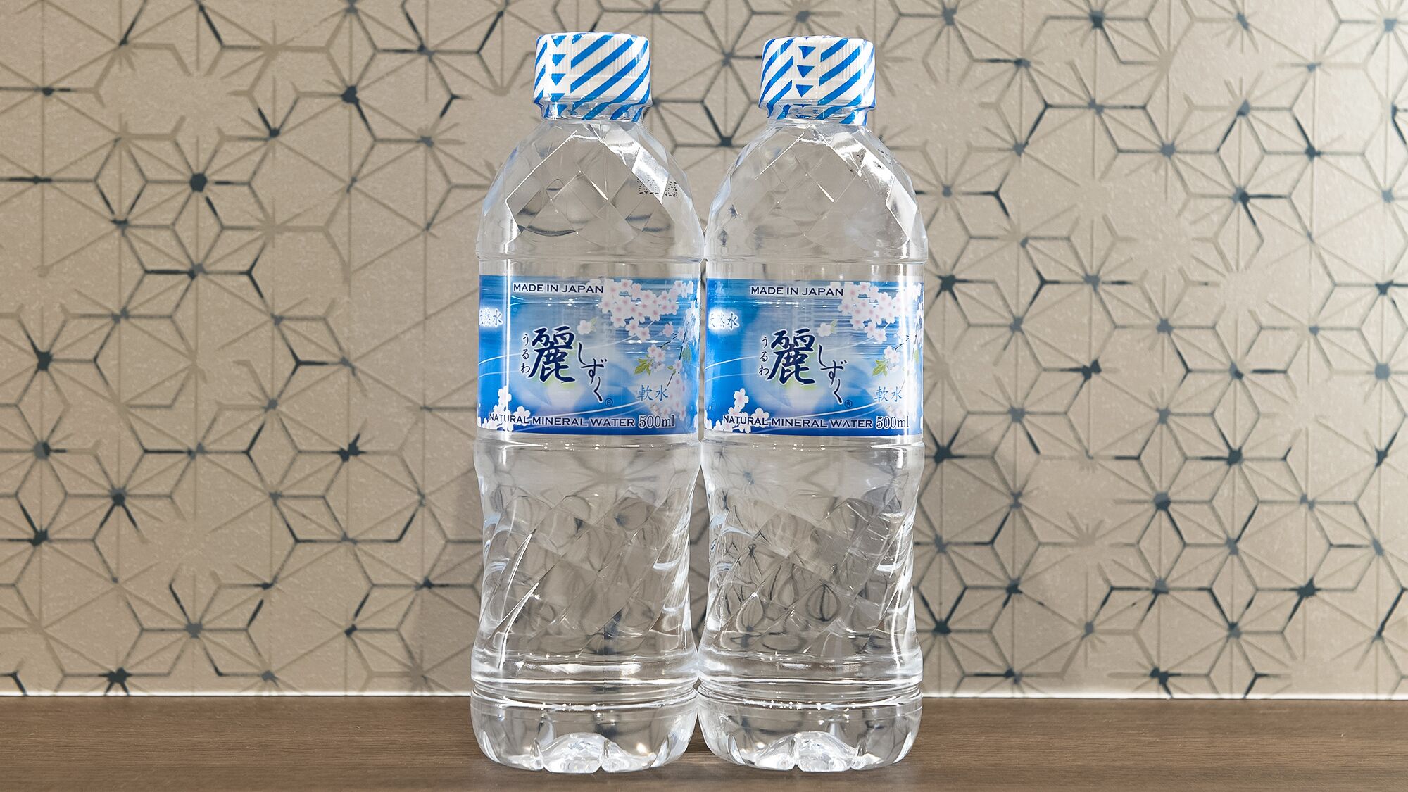 Two clear plastic water bottles with blue and white labels, placed on a wooden surface against a geometric patterned wall.