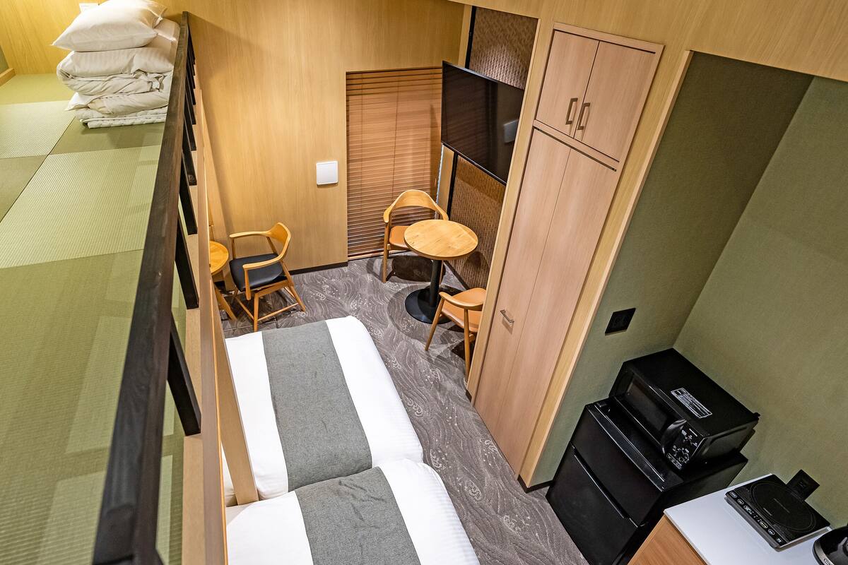 A compact hotel room with tatami mat flooring, a bed with white bedding, a small dining area with a table and chairs, and a kitchenette with a stove.