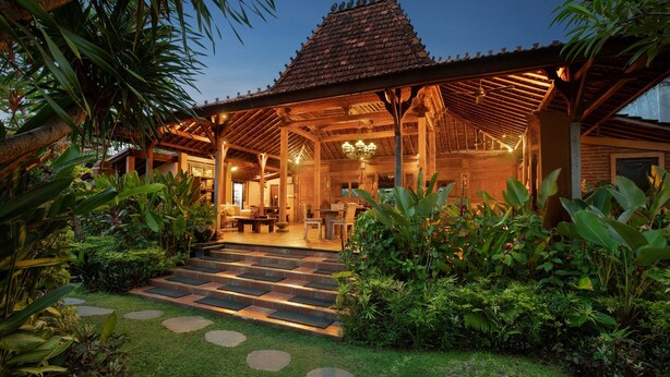 A traditional wooden structure with a tiled roof, surrounded by lush greenery and a stone pathway leading to a seating area. - Mahe Garden Inn and Villas (Kuta)