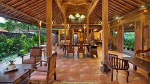 A covered patio with wooden furniture and a tiled floor, surrounded by lush greenery. - Mahe Garden Inn and Villas (Kuta)