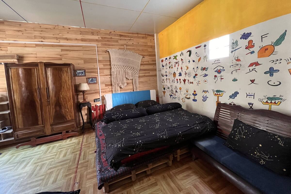 Ein Zimmer mit Holzboden, einem Bett mit gemustertem Überwurf, einem Holzschrank, einem Wandbehang aus Makramee und einer Wand mit verschiedenen Zeichnungen.