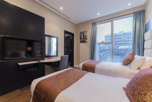 A hotel room with two beds, a desk with a chair, a television, and a large window with a city view. - Hyatt Buyutat Service Apartments (Riyadh)