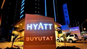 Señal del hotel Hyatt Buyutat con letras iluminadas, fachada de un edificio moderno y un área de estacionamiento con vehículos.