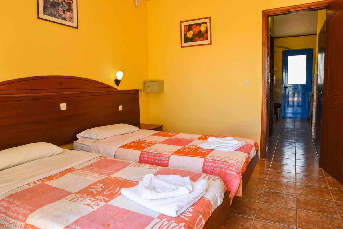 A hotel room with two beds, a wooden headboard, a nightstand, a wall-mounted lamp, and a door leading to a corridor.