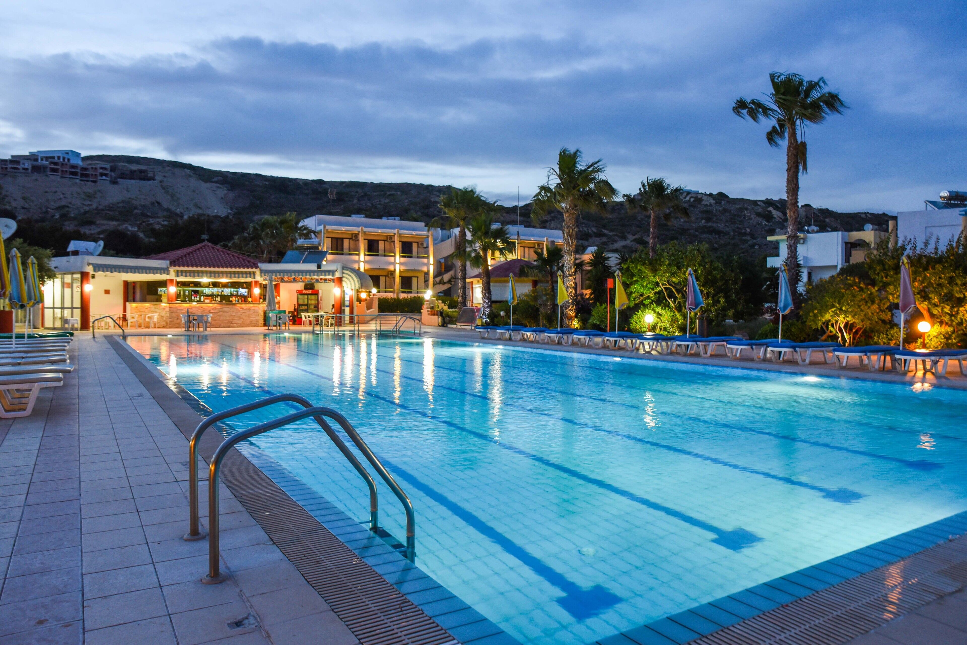 A swimming pool with lounge chairs and umbrellas, surrounded by buildings and palm trees.